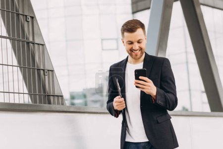 Photo for Portrait of happy modern male businessman in suit walking smiling and reading or writing message on phone on background of urban buildings and offices. - Royalty Free Image