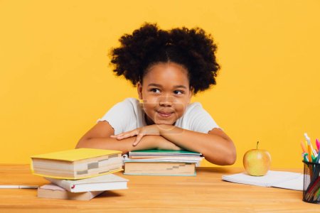 Happy African American schoolgirl sitting at desk leaning on books. Back to school concept. Poster 643666354