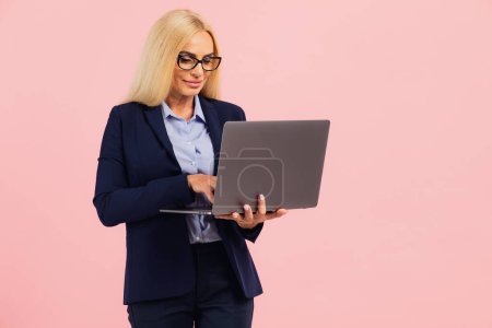 Foto de Atractiva mujer de negocios de mediana edad en gafas con un portátil sobre un fondo rosa - Imagen libre de derechos