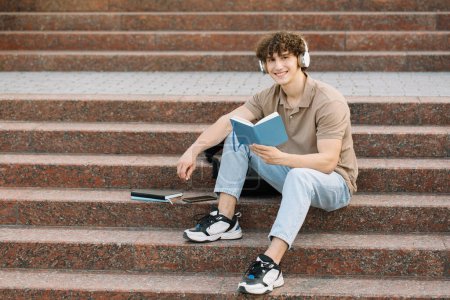 Téléchargez les photos : Étudiant masculin attrayant assis sur les marches de l'université et lit quelque chose, se préparant pour les examens - en image libre de droit
