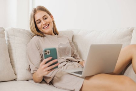 Foto de Hermosa mujer rubia en pijama tumbada en un sofá cerca de la computadora portátil y mirando el teléfono, concepto de trabajo desde casa, cuarentena, nueva normalidad - Imagen libre de derechos