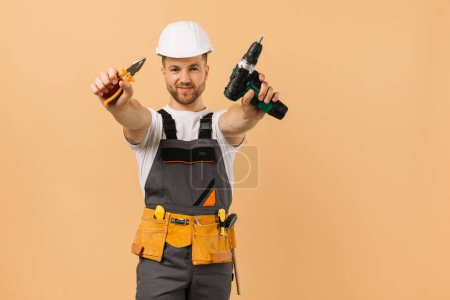 Téléchargez les photos : Réparateur masculin positif à la maison montre tournevis et pinces sur fond beige - en image libre de droit