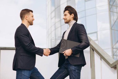 Photo for Two modern businesspeople greet each other smiling and shaking hands against the background of urban offices and buildings - Royalty Free Image