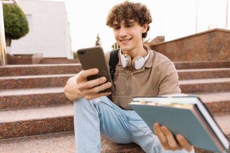 Téléchargez les photos : Attrayant bouclé jeune homme université ou étudiant est assis sur les escaliers et à la recherche smartphone - en image libre de droit