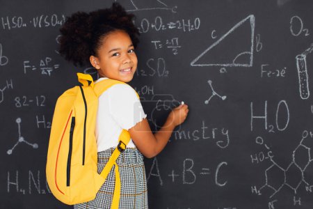 Foto de Feliz colegiala afroamericana resolviendo problemas cerca de la pizarra en la escuela, de vuelta al concepto escolar. - Imagen libre de derechos