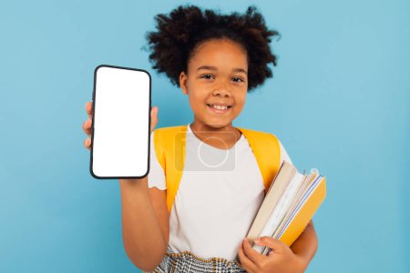 Foto de Emocionado Little African American School Girl sosteniendo una gran celda en la mano presentando la pantalla vacía del teléfono en el fondo del estudio azul. Smartphone Display Mockup - Imagen libre de derechos