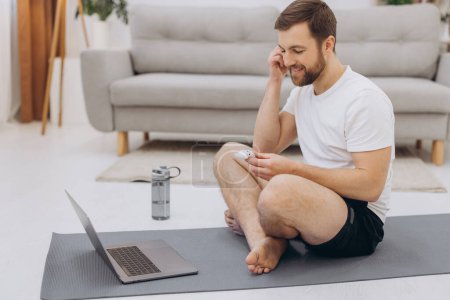 Téléchargez les photos : Homme barbu attrayant tenant des écouteurs sans fil près d'un ordinateur portable et commençant l'exercice du matin ou yoga. - en image libre de droit