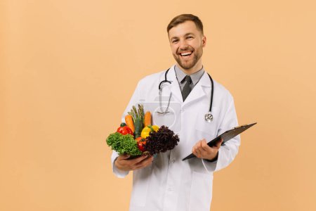 Photo for The happy male doctor nutritionist with stethoscope holding fresh vegetables on beige background, diet plan concept - Royalty Free Image