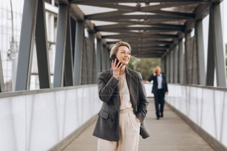 Photo for Portrait of a beautiful mature business woman in suit and gray jacket smiling and talking on the phone on a modern urban and office buildings background - Royalty Free Image