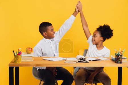 Téléchargez les photos : Heureuse écolière afro-américaine et écolier métis assis ensemble au bureau et se donnant cinq sur fond jaune. Concept de retour à l'école. - en image libre de droit