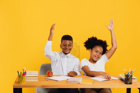 Photo for Happy african american female schoolgirl and mixed race schoolboy sitting together at desk and stretching arms up on yellow background. Back to school concept. - Royalty Free Image
