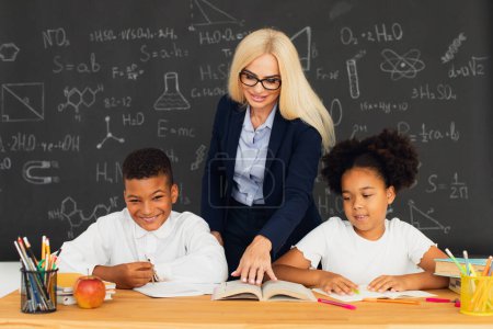 Photo for A female teacher teaches school children who sitting at a desk and solving problems. International team. Back to school. - Royalty Free Image