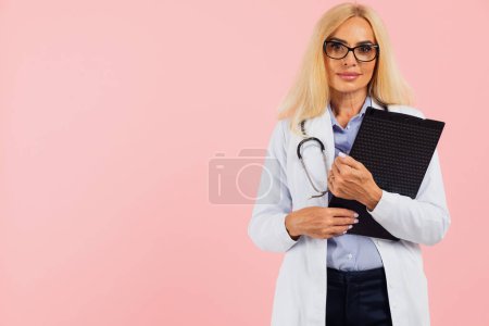 Photo for Mature woman doctor in glasses with stethoscope holding folder on the pink background with copy space - Royalty Free Image
