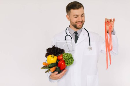 Foto de El médico nutricionista masculino con estetoscopio sosteniendo verduras frescas y regla sobre fondo blanco - Imagen libre de derechos