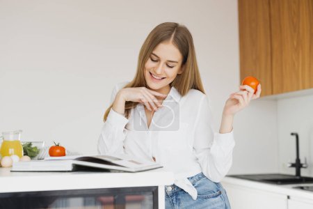 Téléchargez les photos : Femme blonde mignonne tenant tomate et regardant un livre de recettes dans la cuisine - en image libre de droit