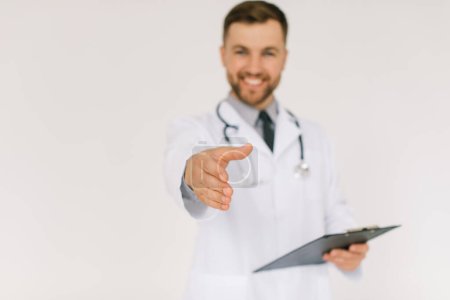 Téléchargez les photos : Le médecin heureux avec un stéthoscope et un dossier étend une main de salutations sur un fond blanc - en image libre de droit