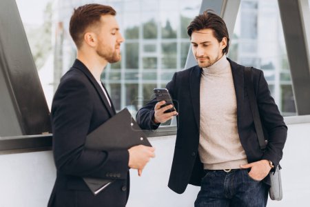 Photo for Two modern happy businessmen with phone and folder of documents discussing something against the background of urban offices and buildings - Royalty Free Image