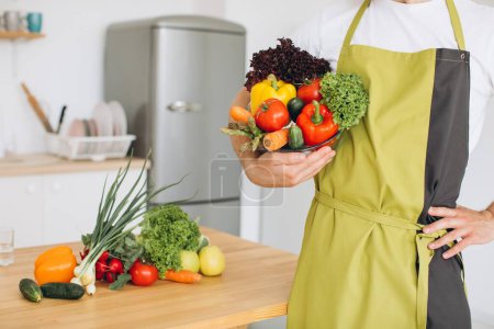 Téléchargez les photos : Fragment d'un homme tenant une assiette de légumes frais sur le fond d'une cuisine et une table de cuisine avec divers légumes. - en image libre de droit
