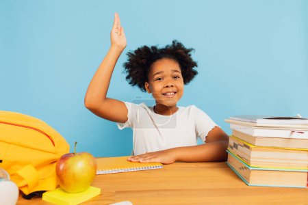 Photo for Happy African American schoolgirl sitting at desk in class and stretching hand uphill on blue background. Back to school concept. - Royalty Free Image