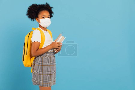 Photo for African american schoolgirl in mask holding books on blue background with copy space. Back to school concept. COVID-19 - Royalty Free Image