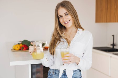 Téléchargez les photos : Une femme blonde mignonne heureuse boit du jus d'orange dans la cuisine - en image libre de droit
