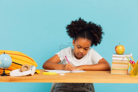 Happy African American schoolgirl doing homework at desk in class on blue background. Back to school concept. tote bag #643670618