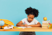 Happy African American schoolgirl doing homework at desk in class on blue background. Back to school concept. Poster #643670618