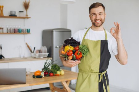 Téléchargez les photos : Portrait d'un homme heureux tenant une assiette de légumes frais sur le fond de la cuisine à la maison - en image libre de droit
