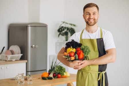 Téléchargez les photos : Portrait d'un homme heureux tenant une assiette de légumes frais sur le fond de la cuisine à la maison - en image libre de droit