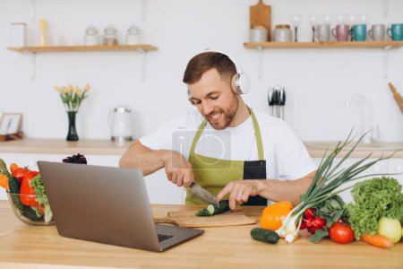 Téléchargez les photos : Homme heureux dans les écouteurs préparer la salade dans la cuisine à partir de légumes frais et en regardant ordinateur portable. - en image libre de droit