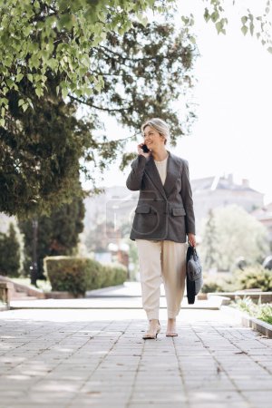 Téléchargez les photos : Mature blonde souriante femme d'affaires marche dans les rues de la ville et parle au téléphone - en image libre de droit
