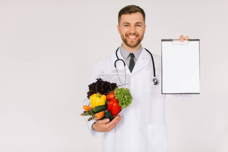 Téléchargez les photos : Le médecin nutritionniste masculin avec stéthoscope tenant des légumes frais et dossier avec espace de copie avec sur fond blanc - en image libre de droit