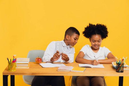 Photo for Happy african american schoolgirl and schoolboy sitting together at desk and studying on yellow background. Back to school concept. - Royalty Free Image