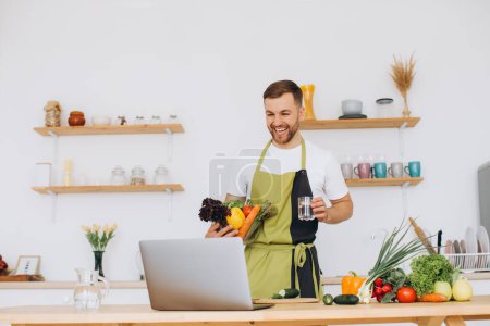 Téléchargez les photos : Portrait de l'homme heureux à la maison, homme cuisine salade de légumes en regardant la caméra et souriant, trancher des légumes, en utilisant un ordinateur portable pour la formation de cuisine en ligne - en image libre de droit