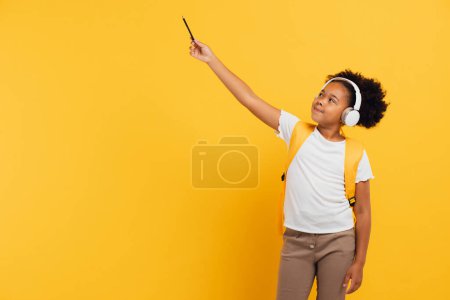 Téléchargez les photos : Heureuse écolière afro-américaine dans un casque avec sac à dos montrant quelque chose avec la main en classe sur fond jaune, espace de copie. - en image libre de droit