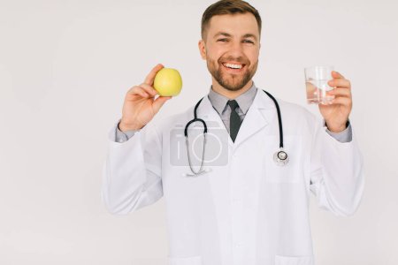Photo for The happy male nutritionist doctor with stethoscope smiling and holding water and apple on white background, diet plan concept - Royalty Free Image
