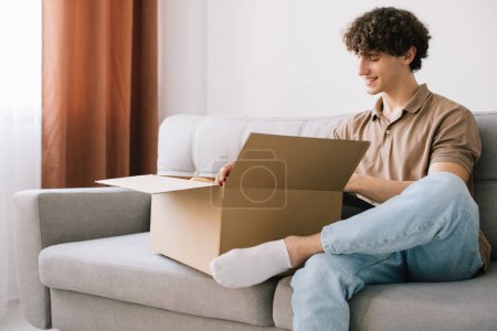 Photo for Happy young smiling curly man opening box with ordered goods gifts, presents at home on couch. Online shopper male customer opening online shop parcel. - Royalty Free Image