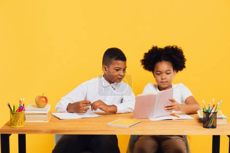 Happy african american schoolgirl and mixed race schoolboy sitting together at desk and studying on yellow background. Back to school concept.