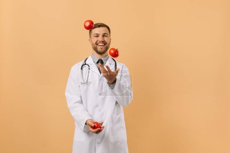 Téléchargez les photos : Le médecin nutritionniste masculin avec stéthoscope souriant et jonglant tomates sur fond beige, concept de plan de régime - en image libre de droit