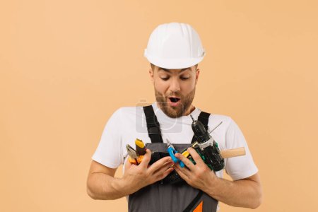 Téléchargez les photos : Réparateur masculin positif à la maison tenant divers outils de réparation sur fond beige - en image libre de droit