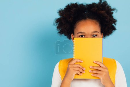 Foto de Divertida colegiala afroamericana cubriéndose la cara con libro amarillo sobre fondo azul, concepto de vuelta a la escuela. - Imagen libre de derechos