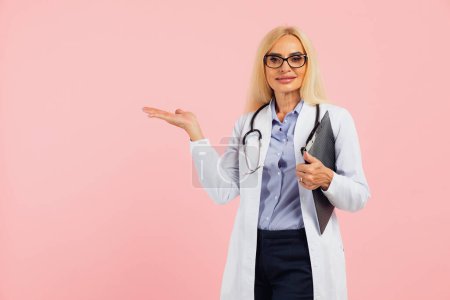 Photo for Mature woman doctor in glasses with stethoscope holding folder and show hand something on copy space the pink background - Royalty Free Image