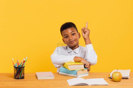Photo for Happy African schoolboy sitting at desk and stretching arm up during class. Back to school concept. - Royalty Free Image