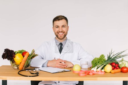 Photo for Happy doctor nutritionist sitting at workplace at desk in office among fresh vegetables, diet plan concept - Royalty Free Image