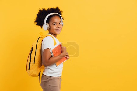 Téléchargez les photos : Heureuse écolière afro-américaine dans un casque avec sac à dos tenant des cahiers sur fond jaune, espace de copie. - en image libre de droit
