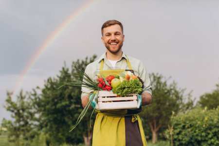 Foto de Hombre jardinero feliz celebración cesta con verduras frescas sobre el arco iris y el fondo del jardín, concepto de jardinería - Imagen libre de derechos