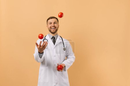 Foto de El médico nutricionista masculino con estetoscopio sonriendo y malabares tomates en fondo beige, concepto de plan de dieta - Imagen libre de derechos