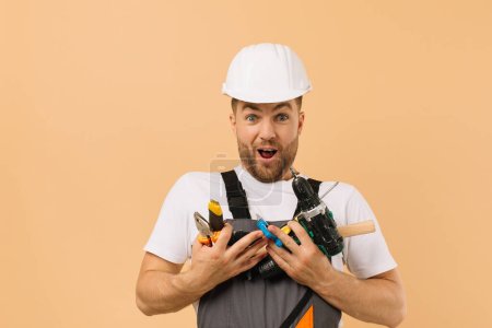 Foto de Reparador masculino positivo en casa sosteniendo varias herramientas de reparación sobre fondo beige - Imagen libre de derechos