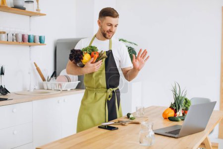 Foto de Retrato del hombre feliz en casa, cocinando ensalada de verduras mirando a la cámara y sonriendo, hace un gesto de saludo con la mano, utilizando el ordenador portátil para el entrenamiento de cocina en línea - Imagen libre de derechos