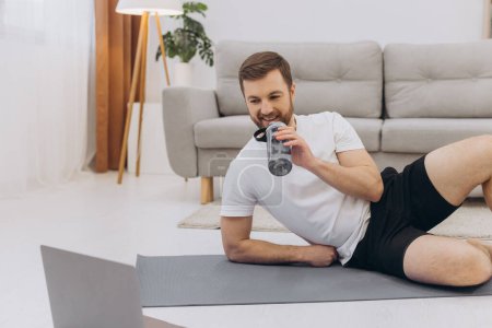 Photo for Handsome adult man using laptop and drinking water while having break during workout at home - Royalty Free Image